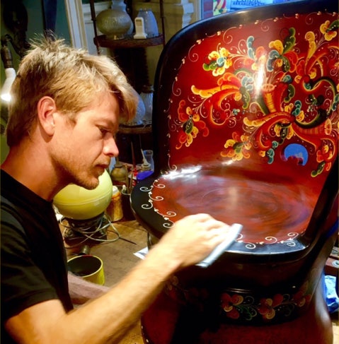 A craftsman hand painting a restored wooden chair with decorative folk art
