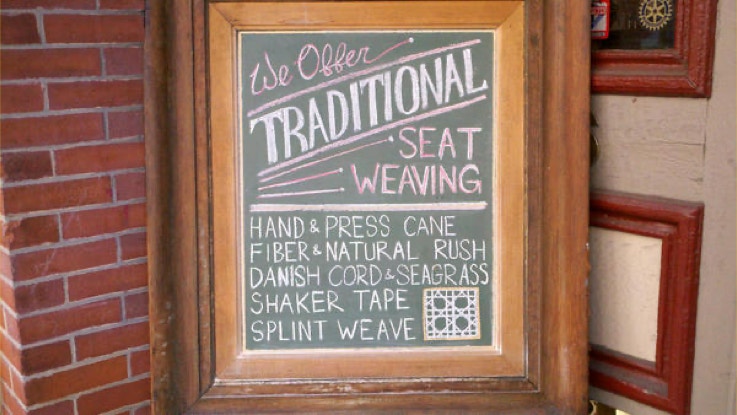 Chalkboard sign in a wood frame listing traditional seat weaving services including hand and press cane, rush, Danish cord, Shaker tape and splint weave
