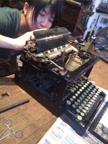 Restorer leaning over an antique black L.C. Smith Model 8 typewriter on a workbench, installing replacement linkages