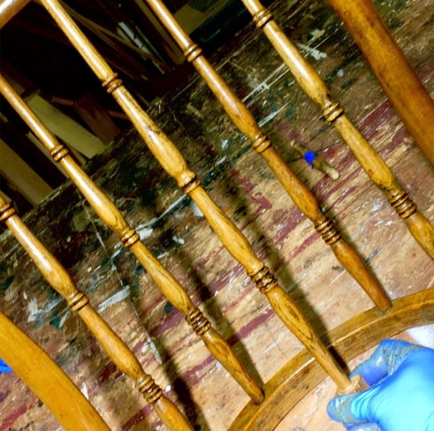Gloved hand checking the color of a newly turned spindle against original spindles in a bow-back rocking chair