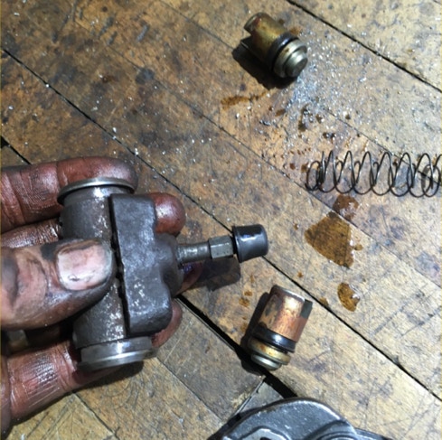 Greasy hand holding a disassembled brake caliper with pistons and spring on a shop floor