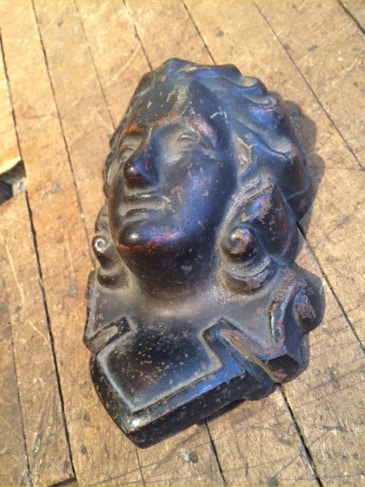 Ornate cast metal sculpture of a face with flowing hair resting on a wooden workshop floor