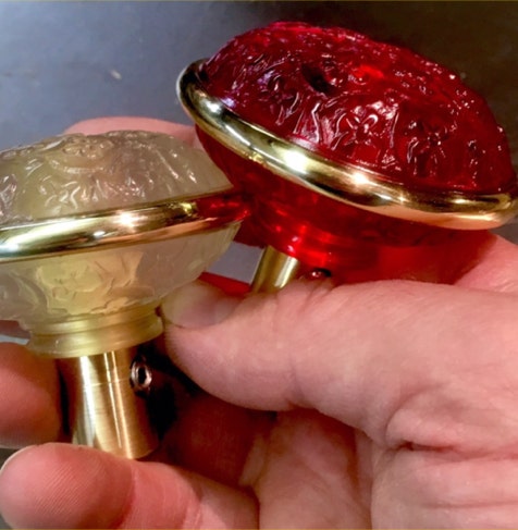 Hand holding two cast resin Madras pattern doorknobs, one cranberry and one frosted, with brass collars