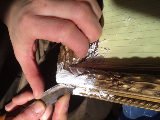 Hands carving leaf detail back into the fresh plaster of a gilt picture frame rail with a small blade