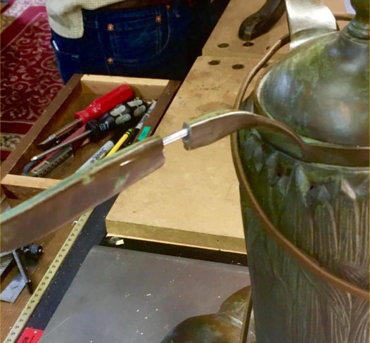 Broken metal arm of a patinated Tiffany-style lamp on a bench with a stainless steel pin being fitted into the break