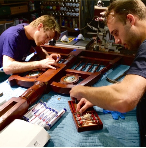 Two restorers working side by side on a mahogany swing bed frame and an antique hydrometer instrument cabinet