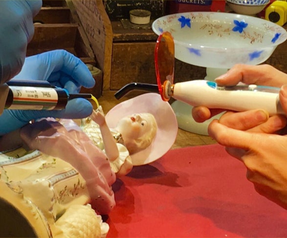 Gloved hands applying light-cured dental composite to the hand of a porcelain figurine while a UV curing light is held nearby