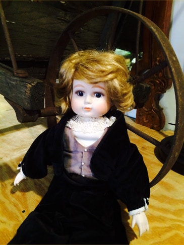 Antique bisque doll with blonde curls in a black velvet dress seated in the workshop