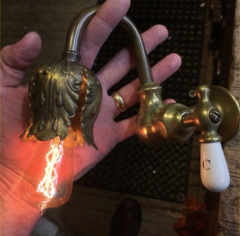 Hand holding a wall sconce built from an antique brass faucet with a glowing filament bulb