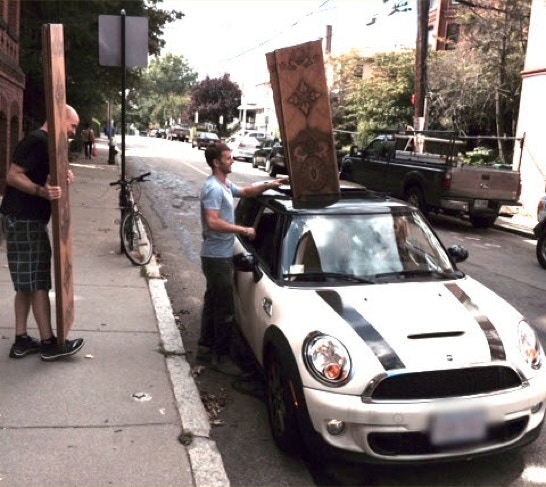 Two people loading tall carved wooden panels through the open sunroof of a Mini Cooper on a city street