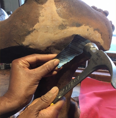Hands texturing an epoxy repair on the shoulder of a cast iron bust using a hammer and coarse sandpaper