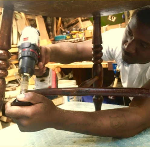 Jordan working underneath an antique chair with a drill, fitting a replacement stretcher in place