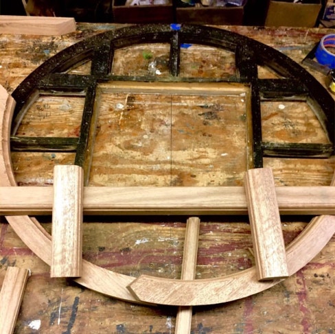 Original arched gable window frame beside newly milled replacement sections on a workbench